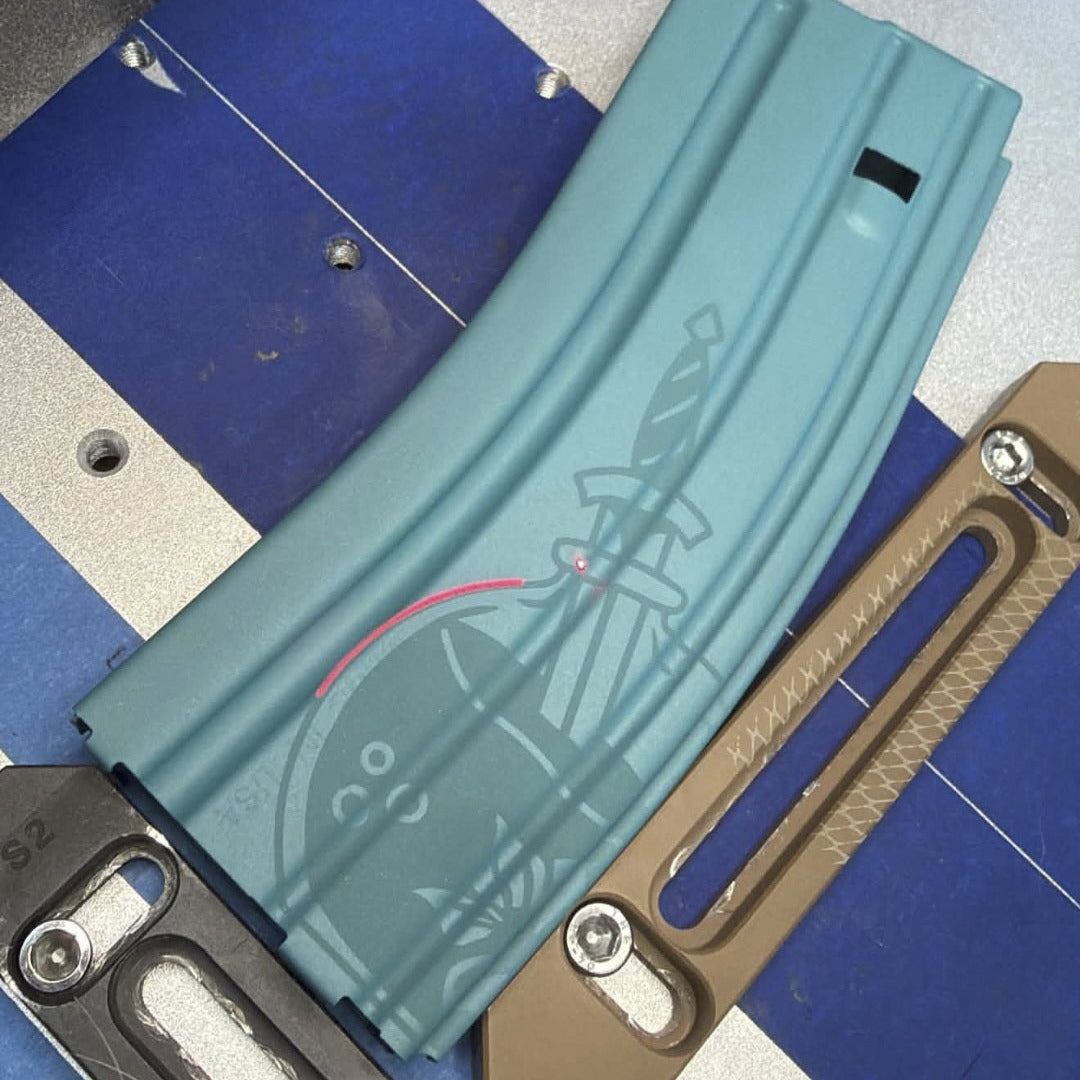 A curved teal tactical magazine or grip component with a sword graphic printed on it, shown alongside two metal clips or tools in black and tan on a blue-and-silver surface.